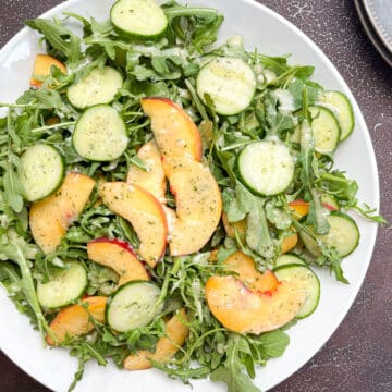 Peach arugula salad with dressing in large white bowl, on top of a back drop next to saucers that are stacked on top of each other.
