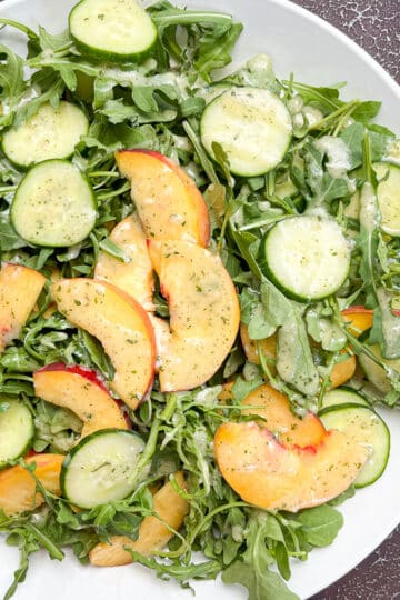 Peach arugula salad with dressing in large white bowl, on top of a back drop next to saucers that are stacked on top of each other.