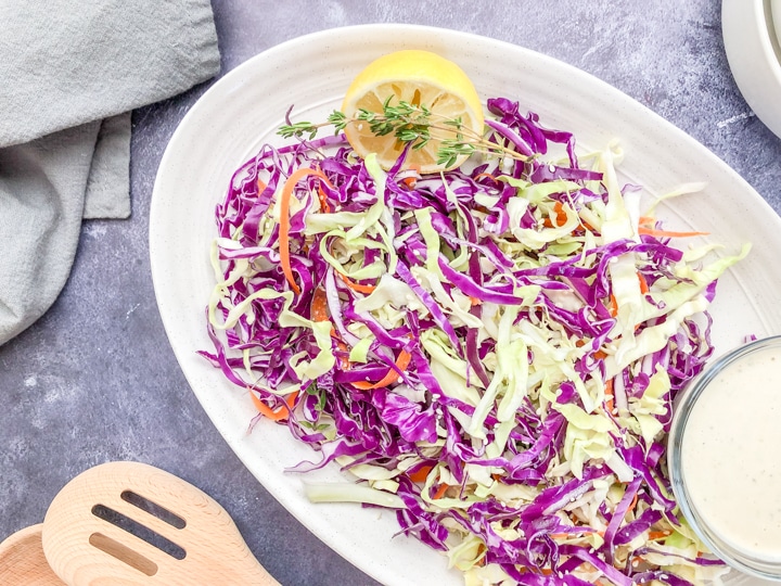 close-up shot healthy coleslaw recipe no mayo in a cream colored oval serving bowl with a side of tahini dressing