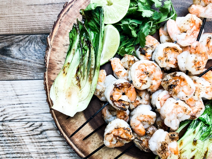 shrimp skewers on round platter garnished with cilantro limes and side of grilled bok choy