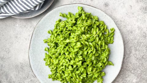 pesto pasta in large light gray plate next to smaller gray plate that has a striped gray napkin on top of it.