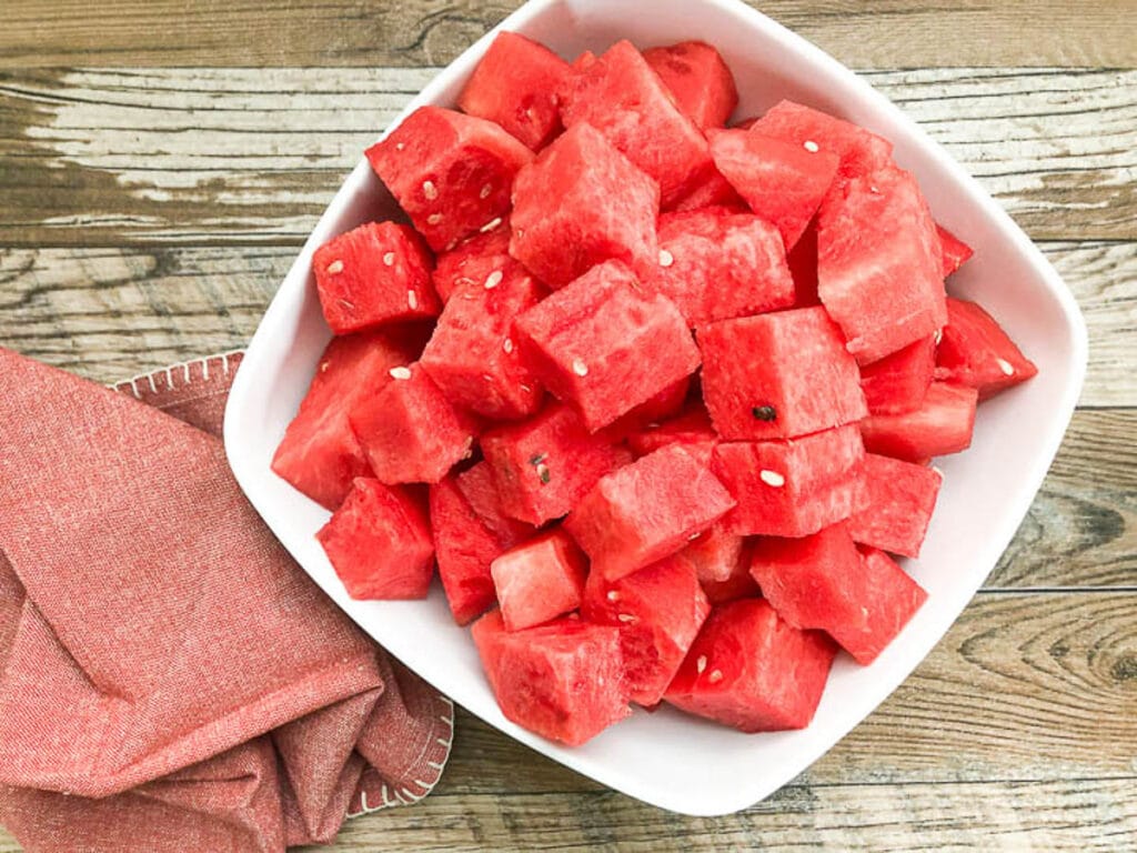 close up of watermelon in white bowl cut into cubes next to red napkin