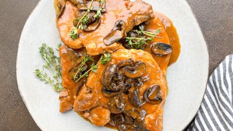Four stacked pork chops with gravy and mushrooms on a gray plate with fresh thyme on the side of the plate and top of the chops.