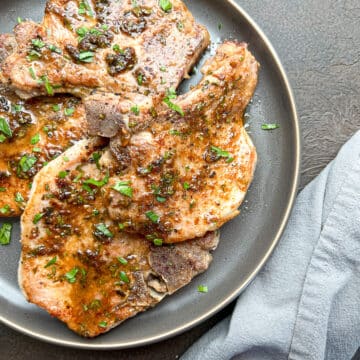 Four lemon pepper pork chops garnished with chopped fresh parsley on a bluish-gray plate next to a gray napkin.
