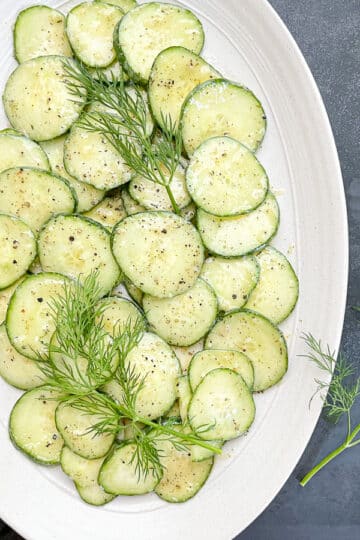 Italian cucumber salad in a white oval serving dish garnished with fresh dill with dill also on the side.