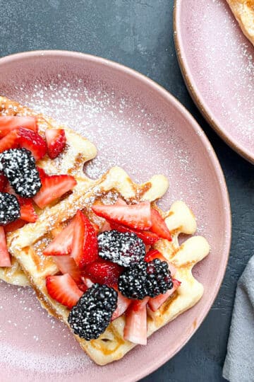 Close-up of waffles topped with berries, dusted with powdered sugar with a drizzle of honey on a pink plate.