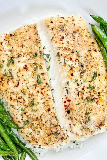 Seasoned baked halibut on a white plate on top of a bed of rice with green beans on the side.