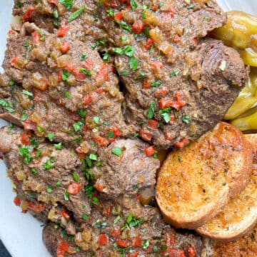 Close-up shot of Mississippi pot roast on a white serving platter with toasted garlic bread and pepperoncini peppers.