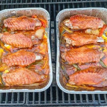 Raw turkey wings, seasoned and placed in aluminum foil cake pans, surrounded by slices of yellow, orange, and green bell peppers and onions.