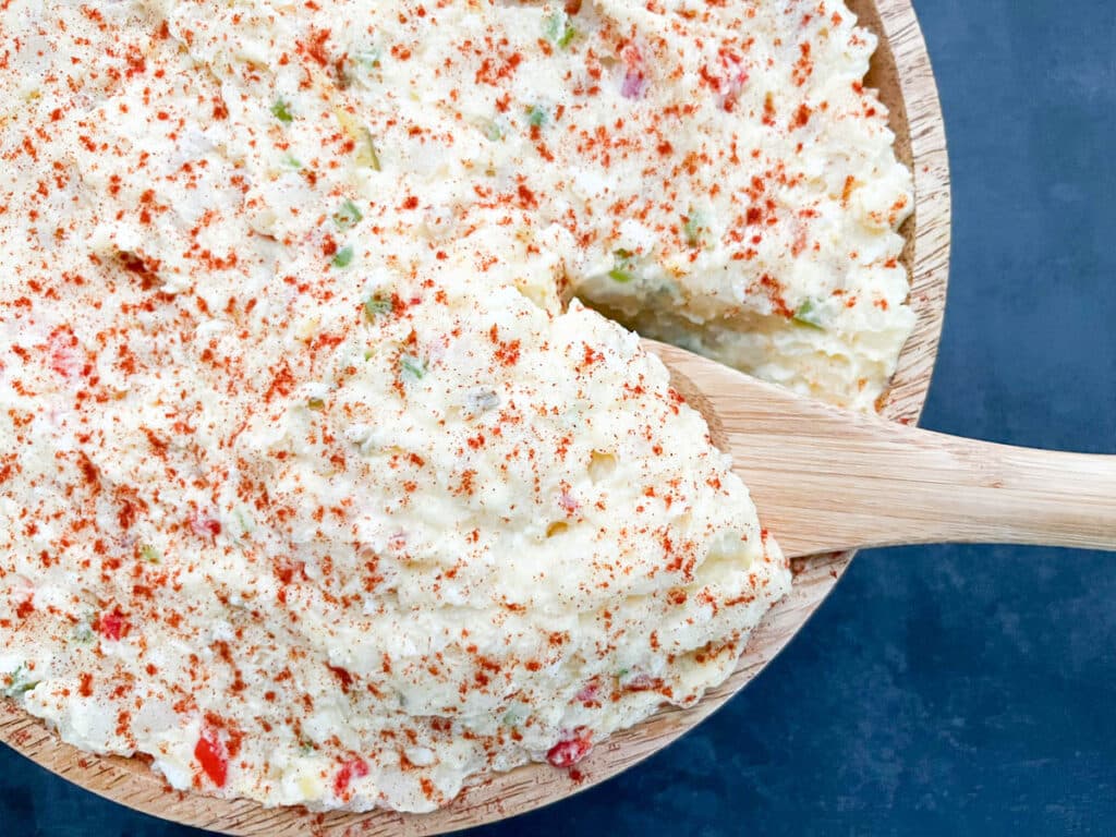 Southern-style potato salad, sprinkled with smoked paprika, served in a wooden bowl with a portion being lifted by a spoon.