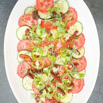 Overhead shot of Southern cucumber and tomato salad, topped with sun-dried tomatoes, thinly sliced jalapeños, and microgreens, all drizzled with a simple vinaigrette on a white oval serving platter.