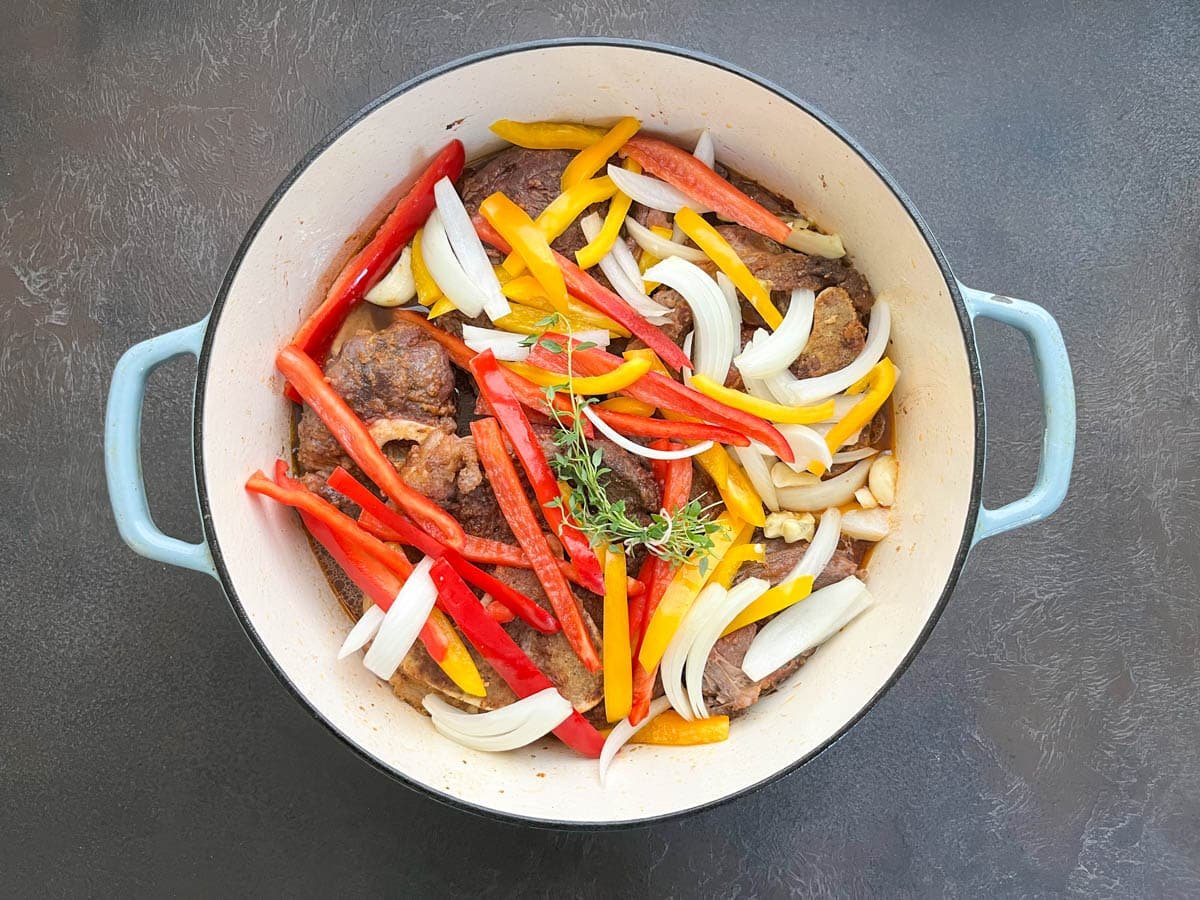 Seared beef shanks in Dutch oven topped with onion, bell peppers, garlic, and fresh thyme before braising in the oven.
