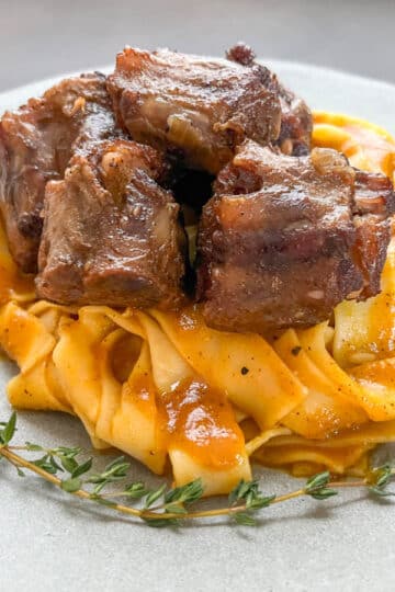 Tender oven-cooked oxtails with rich gravy over pappardelle pasta on a gray plate.
